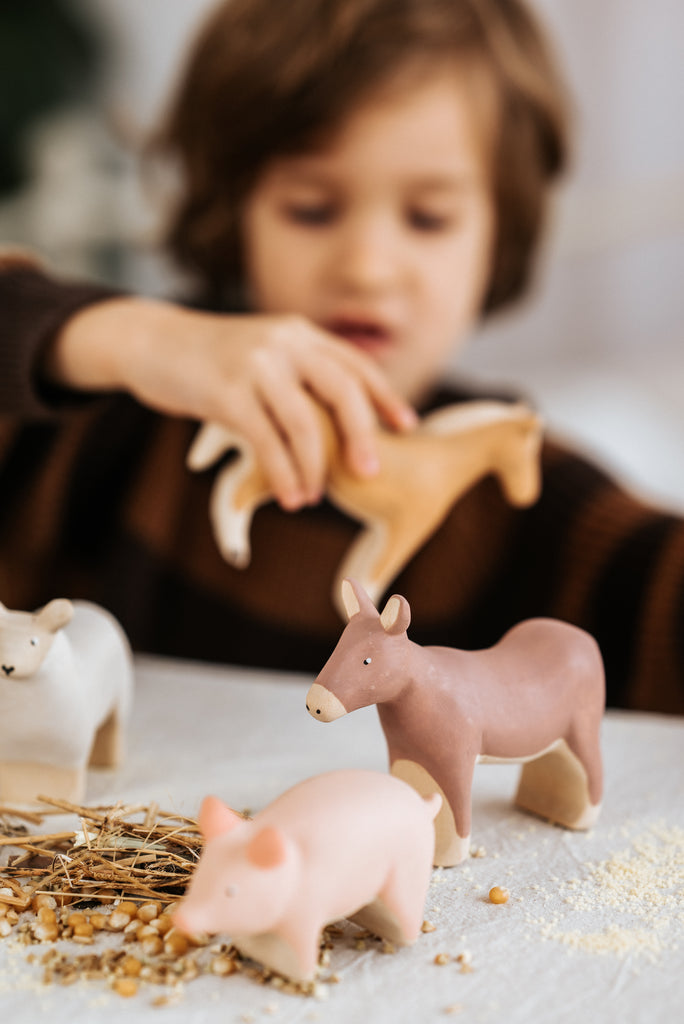 A child enjoys handmade wooden toys—a pink pig, a cow, and a Handmade Wooden Donkey—all crafted from linden wood with child-safe paint. These charming farm animals sit on a hay-covered table as the child plays in the background.