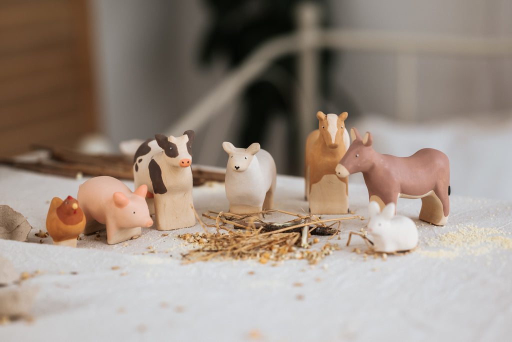 The Handmade Wooden Donkey, crafted from linden wood, is displayed on a bed with scattered straw and seeds among figurines of a cow, sheep, goat, pig, chicken, and rabbit. A softly blurred background using child-safe paint adds to the cozy atmosphere.