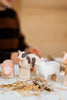 A Handmade Wooden Sheep figurine, crafted from linden wood, sits on a straw-scattered surface alongside farm animals. A person in a brown and black striped shirt is partially visible in the background.