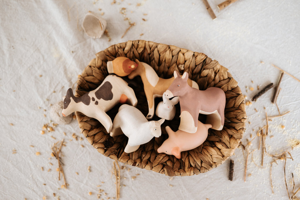A woven basket holds six small, linden wood farm animals painted with child-safe paint. The whimsical set features a cow, pig, horse, dog, sheep (Handmade Wooden Sheep), and chicken on a textured surface amid scattered straw and twigs.