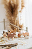 The Handmade Wooden Donkey, crafted from linden wood with child-safe paint, adds a rustic touch when displayed with farm animal figurines like a cow, horse, dog, sheep, and pig on a white tablecloth amid scattered straw and tall dried pampas grass.