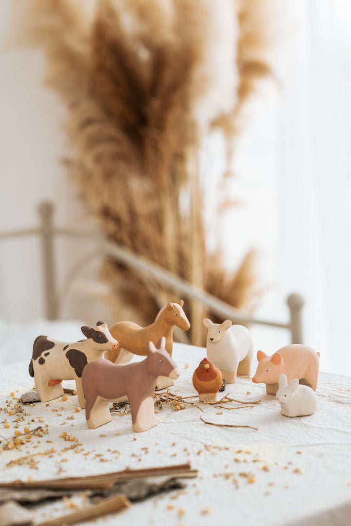 The Handmade Wooden Donkey, crafted from linden wood with child-safe paint, adds a rustic touch when displayed with farm animal figurines like a cow, horse, dog, sheep, and pig on a white tablecloth amid scattered straw and tall dried pampas grass.