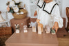 A child in a white dress plays with the Handmade Wooden Hippo and other wooden toys like a zebra, elephant, and giraffe. The vibrant figurines, coated with child-safe paint, sit on a table. A wicker basket and flowers add charm to the serene scene in the background.