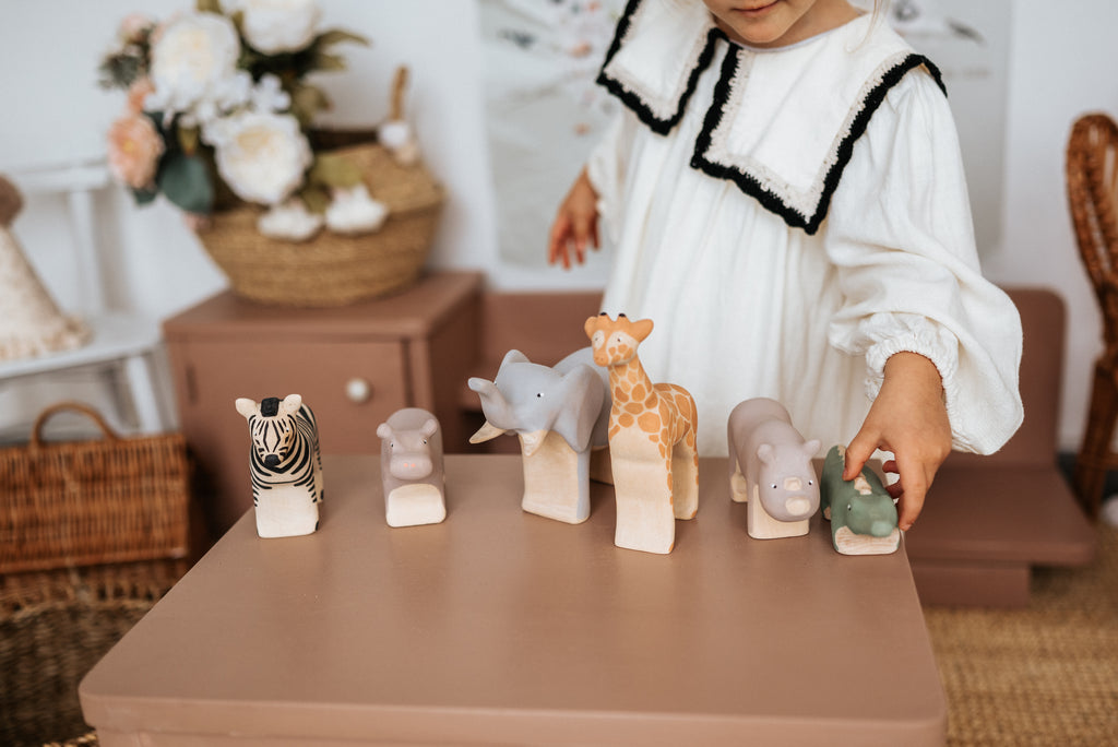 A child in a white dress plays with the Handmade Wooden Hippo and other wooden toys like a zebra, elephant, and giraffe. The vibrant figurines, coated with child-safe paint, sit on a table. A wicker basket and flowers add charm to the serene scene in the background.