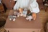 A child in a white shirt joyfully plays with handcrafted wooden animals, including a zebra, elephant, and the Handmade Wooden Hippo on a brown table. Each toy features child-safe paint. Cozy charm is added by wicker baskets and a woven rug in the background.