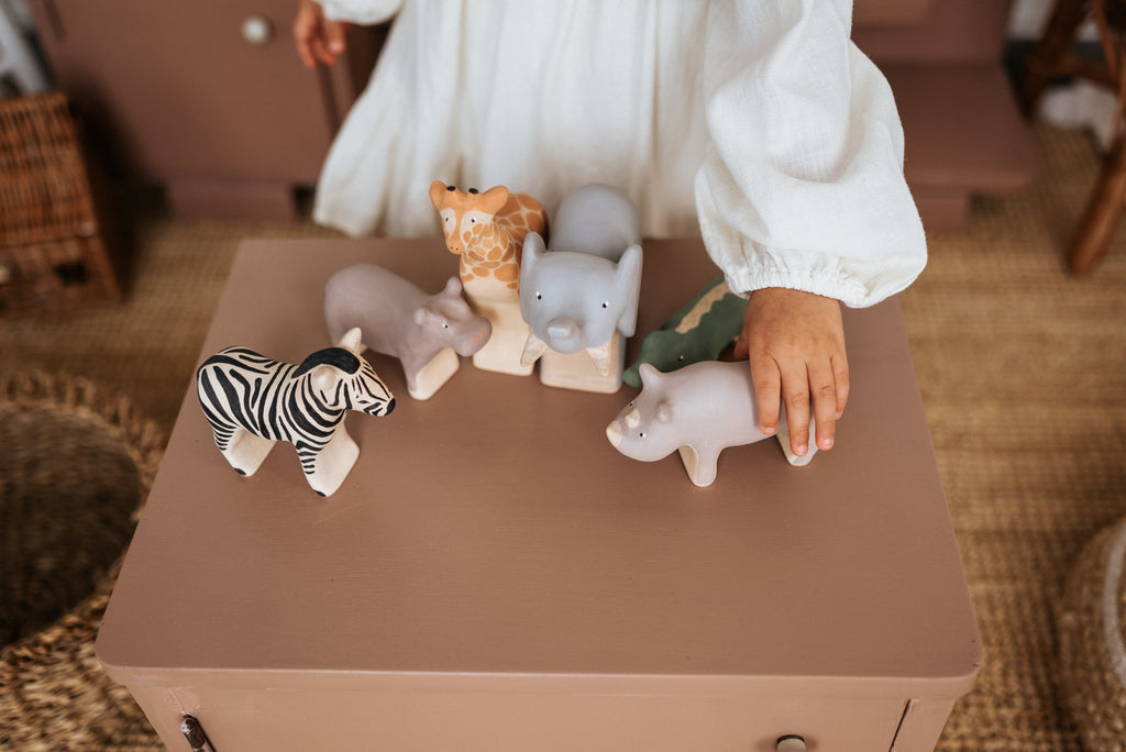 A child in a white shirt joyfully plays with handcrafted wooden animals, including a zebra, elephant, and the Handmade Wooden Hippo on a brown table. Each toy features child-safe paint. Cozy charm is added by wicker baskets and a woven rug in the background.
