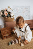 A child in a white dress plays with a Handmade Wooden Hippo on a woven rug. Behind her, there's a wicker basket, pale pink sideboard, and floral arrangement. A pastel wall art piece completes the serene scene.
