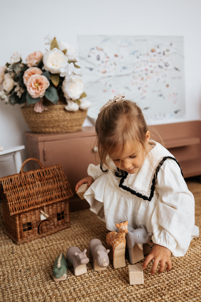 A child in a white dress plays with a Handmade Wooden Hippo on a woven rug. Behind her, there's a wicker basket, pale pink sideboard, and floral arrangement. A pastel wall art piece completes the serene scene.