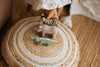 A person arranges a Handmade Wooden Hippo alongside a zebra and crocodile on a round, woven rug. Each toy is crafted with child-safe paint and detailed meticulously. The individual wears white, with a fuzzy white object partially visible in the background.