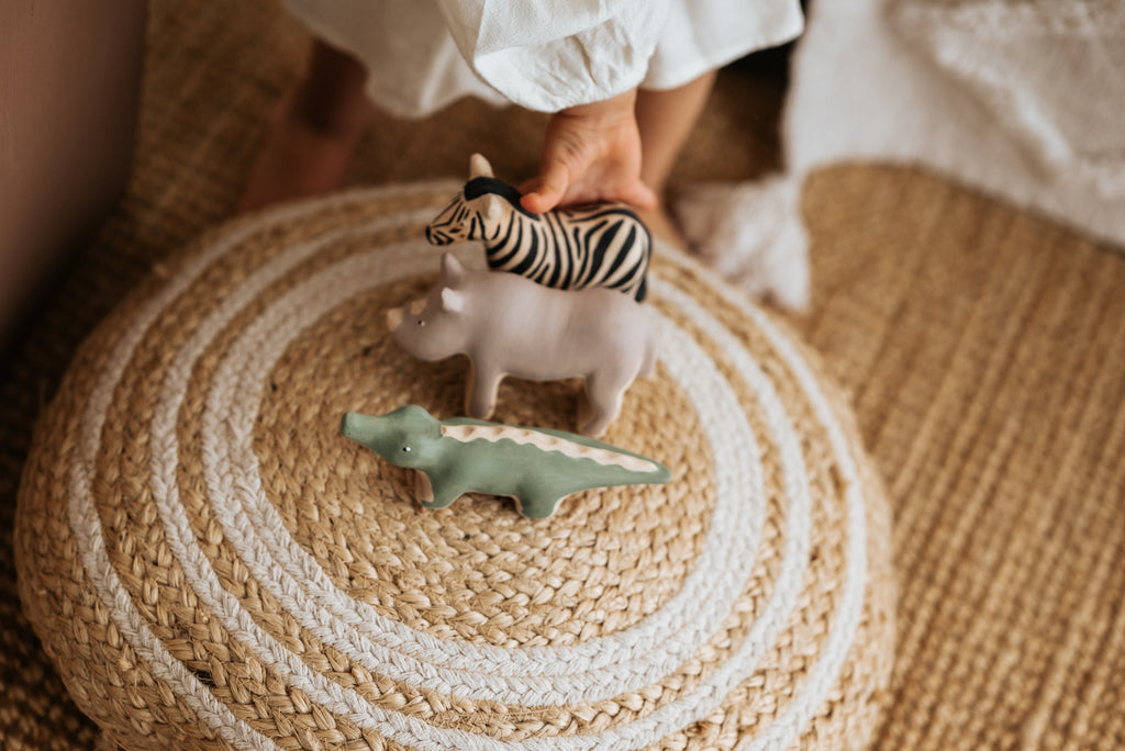 A person arranges a Handmade Wooden Hippo alongside a zebra and crocodile on a round, woven rug. Each toy is crafted with child-safe paint and detailed meticulously. The individual wears white, with a fuzzy white object partially visible in the background.