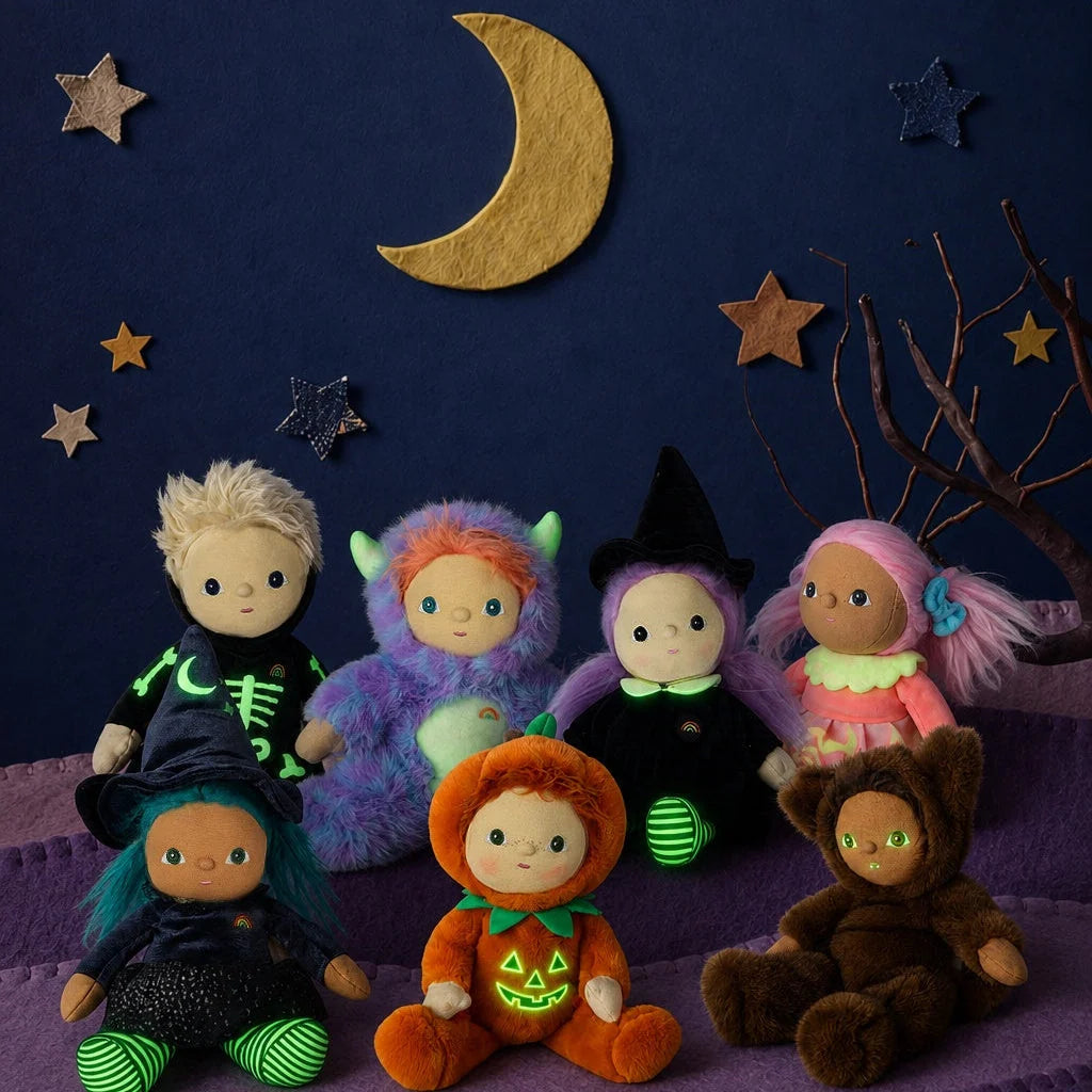 Seven small Halloween stuffed animal dolls, including an Olli Ella Dinky Dinkum Doll - Miles Monster, a pumpkin, and witches, arranged on a purple blanket against a backdrop with a moon and stars.