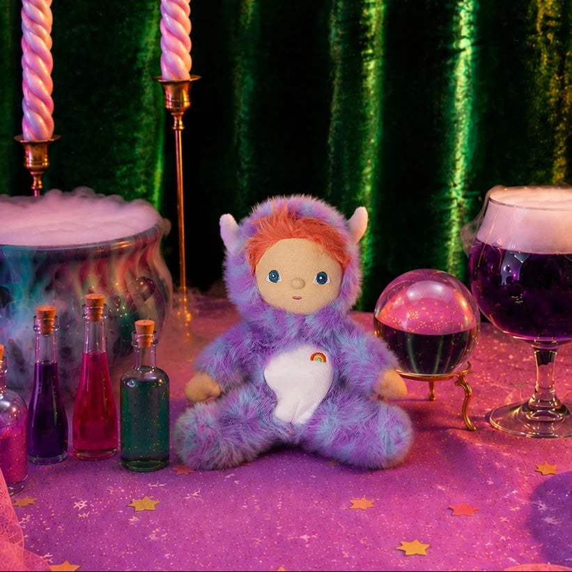 An Olli Ella Dinky Dinkum Doll - Miles Monster with orange hair and a purple and blue fuzzy suit, featuring a white belly patch with a small rainbow, sits on a purple table surrounded by magic potion bottles, a crystal ball, and a bubbling cauldron.