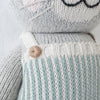 Close-up of Cuddle + Kind Dylan the Kitten, a hand-knit gray cat plush in a light blue and white striped outfit with a brown button on the strap; this stuffed toy features embroidered whiskers and mouth, perfect for snuggles.