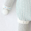 A close-up of the Cuddle + Kind Dylan the Kitten hand-knit doll, featuring grey and white arms, a white hand, and a blue and white vertically striped body on a soft, textured white background.