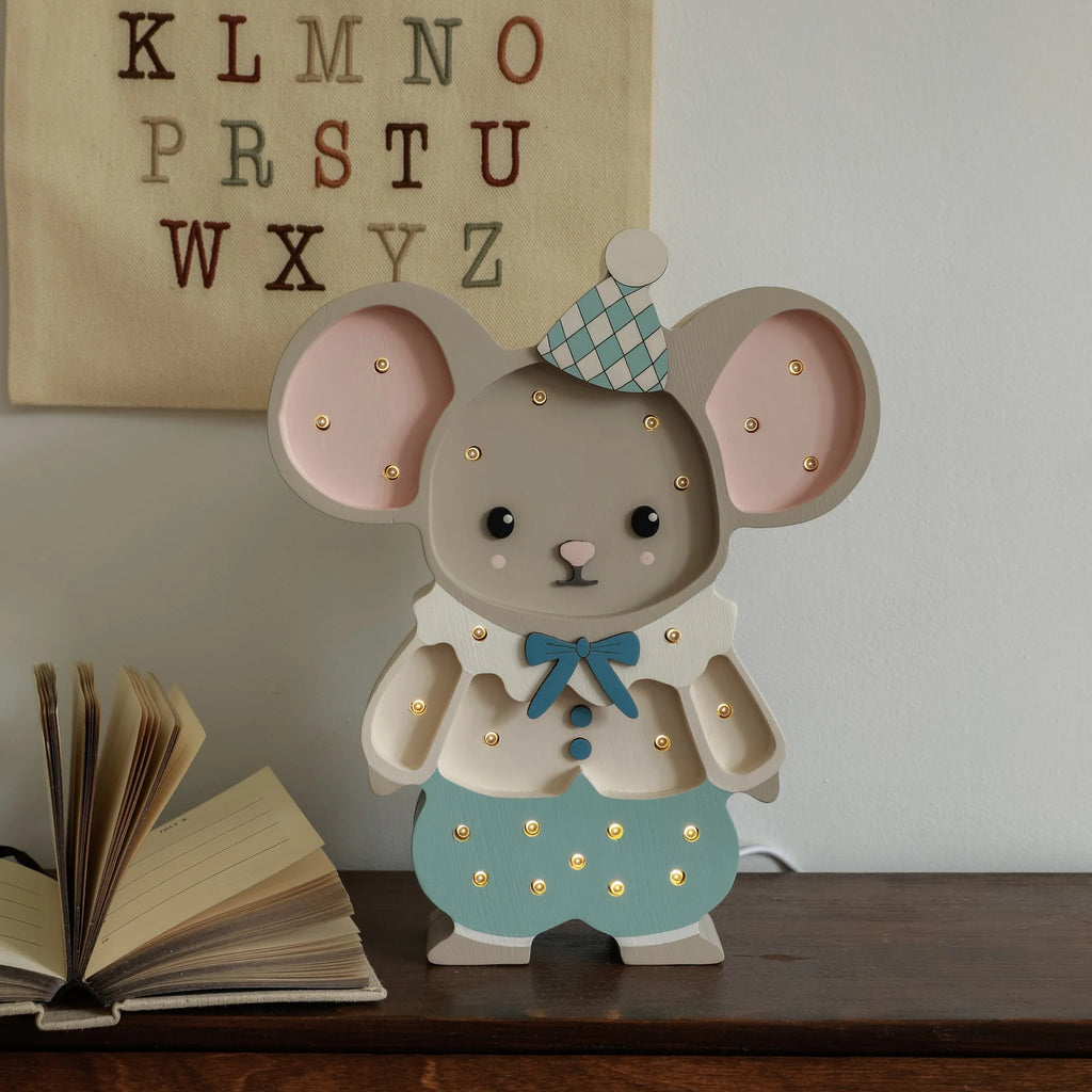 The Handmade Wooden Lamp - Felix The Mouse, featuring a mouse in blue and white clown clothes, stands on a wooden surface by an open book with an alphabet wall hanging—an adorable addition to any nursery decor.