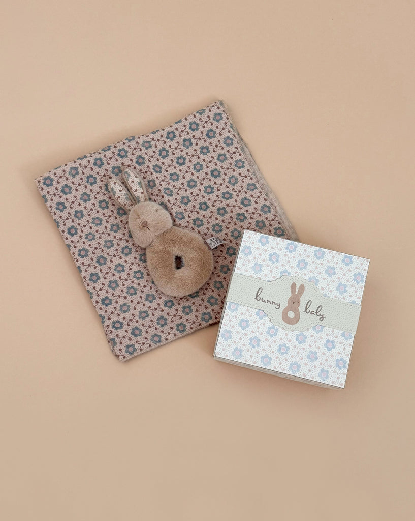 A Newborn Gift Set featuring a soft, floral-patterned bunny rattle and burp cloth with a plush bunny toy is displayed beside a pastel "bunny baby" gift box on a beige background.