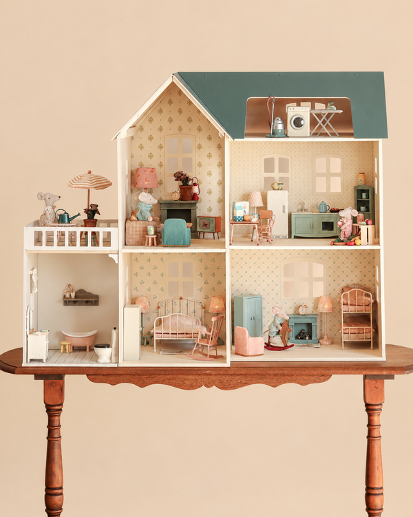 The Maileg Fully Furnished Dollhouse Set + Surprise Gift is displayed on a wooden table, showcasing intricately detailed miniature furniture, lamps, and animal figurines arranged in different rooms—such as the kitchen, bathroom, bedroom, and balcony.