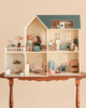 The Maileg Fully Furnished Dollhouse Set + Surprise Gift is displayed on a wooden table, showcasing intricately detailed miniature furniture, lamps, and animal figurines arranged in different rooms—such as the kitchen, bathroom, bedroom, and balcony.