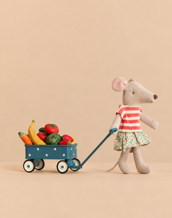 A Maileg mouse in a striped shirt and floral skirt pulls a green Maileg Grocery Wagon Set filled with miniature toy vegetables.