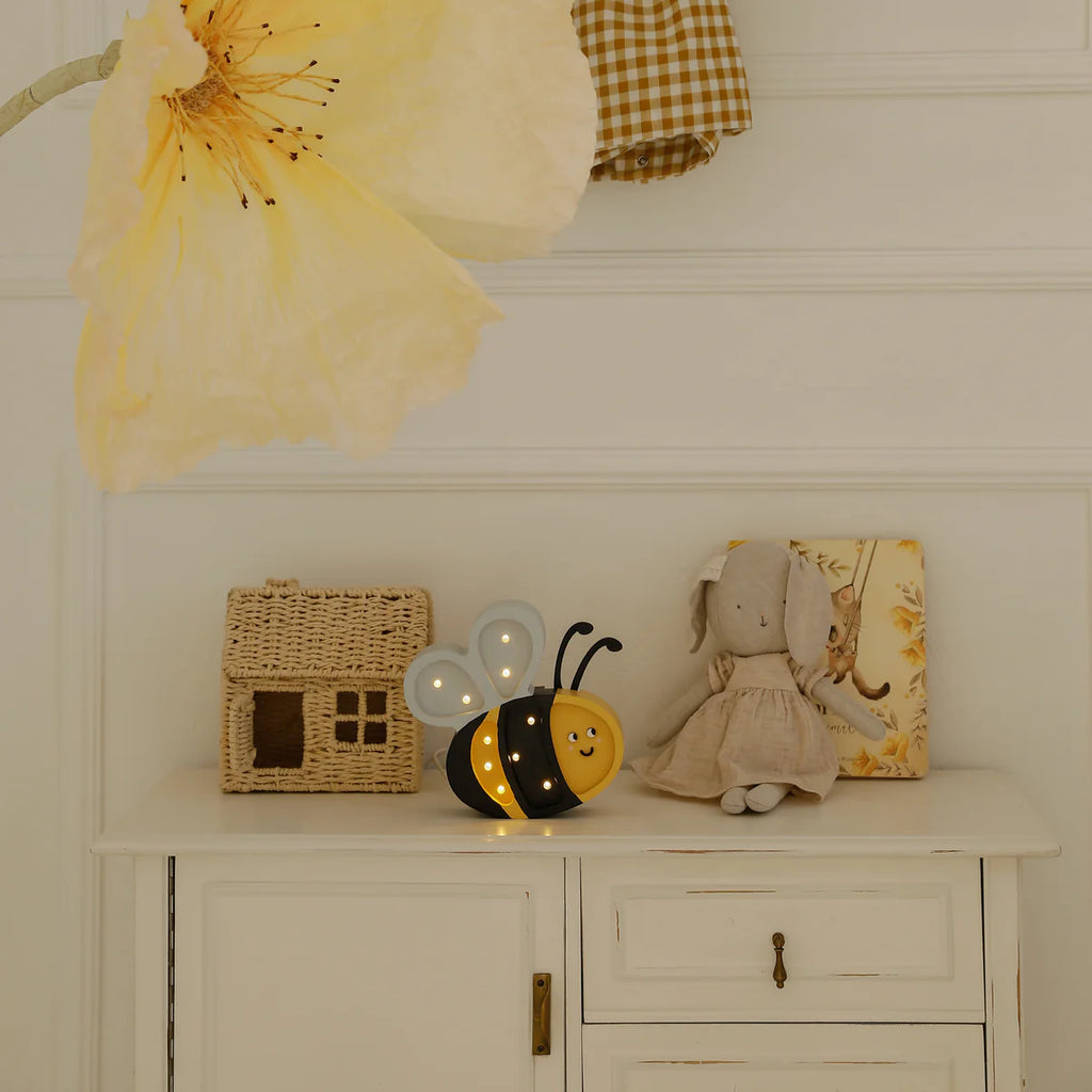 A white cabinet displays a wicker house, the Handmade Wooden Mini Lamp - Happy Bee, and a stuffed animal in a dress, with a plaid fabric and big yellow flower hanging above—an adorable setup for kids' room decor.