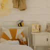 A cozy kids' room features a bed with a sunburst pillow, a doll, the Handmade Wooden Mini Lamp - Happy Bee as a whimsical night light, a gingham dress, the book "Who am I?", a bunny plush, and a white nightstand.