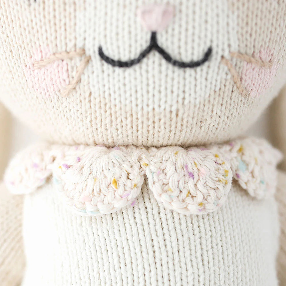 Close-up of Cuddle + Kind Hannah the Bunny, a hand-knit doll made from premium cotton yarn, with embroidered black mouth, pink nose, whiskers, and a scalloped collar adorned with pastel specks.