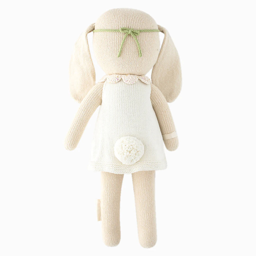 Cuddle + Kind Hannah the Bunny is a beige hand-knit doll made from premium cotton yarn, featuring a white dress, fluffy tail, and green headband, shown from the back on a white background.