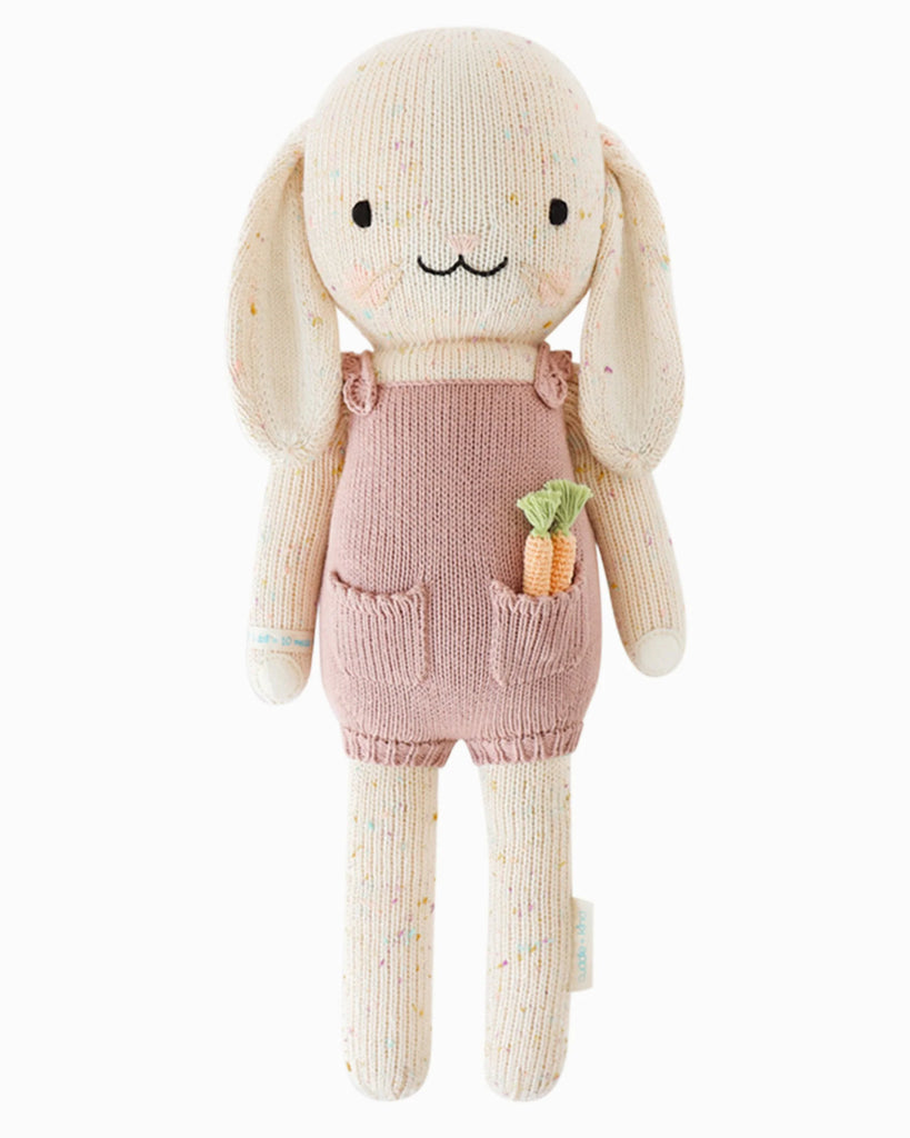 Cuddle + Kind Harper the Bunny is a hand-knit doll from premium cotton, featuring long floppy ears, a happy face, and pink overalls with a carrot in the pocket. Handmade in Peru.