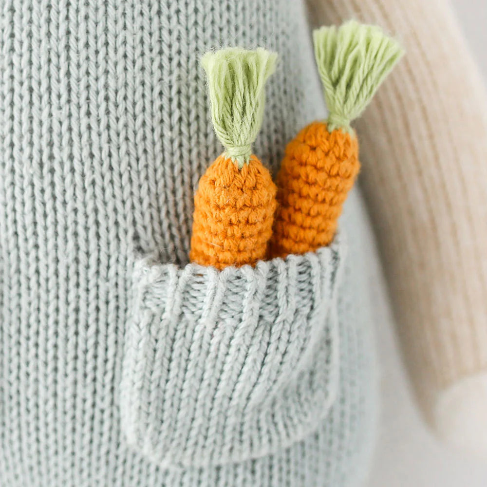 Close-up of two orange crocheted carrots with green tops, made from organic cotton yarn, tucked into the pocket of a light gray Cuddle + Kind Henry the Bunny.