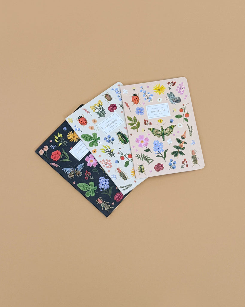 The Assorted Set of 3 Curio Notebooks, each with unique gold foil floral and botanical designs on the cover, is displayed in a fan arrangement on a beige background. Each ruled notebook showcases distinct colorful plant patterns.