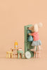 A toy mouse in a striped shirt and skirt stands on the Maileg Stool Set, Mouse - Light Rose/Light Mint, reaching into a green cabinet with a charming vintage paint finish. Small stools from the set, a bottle, and a plate with pastries are nearby, all delicately arranged on a beige background.