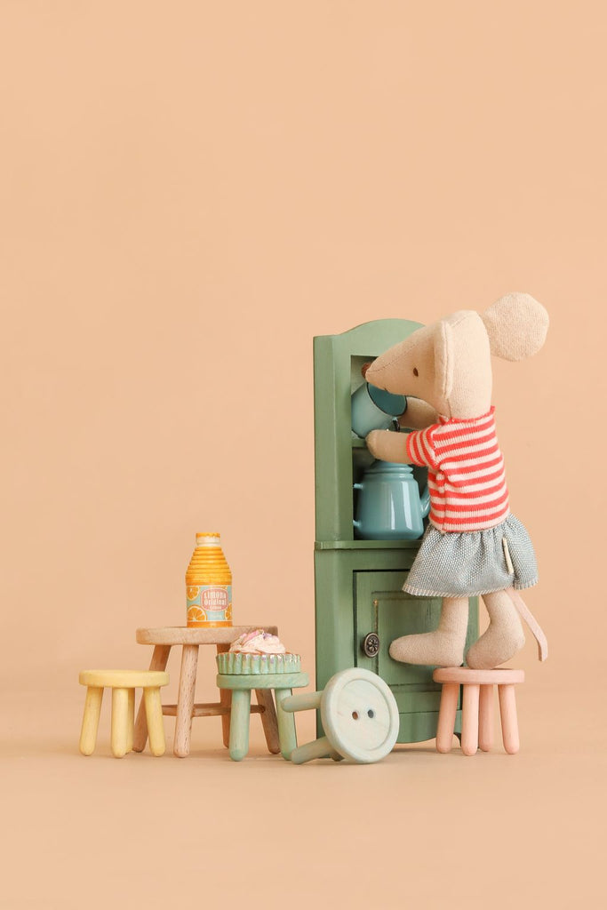 A toy mouse in a red-striped shirt and blue skirt stands on the Maileg Stool Set, Mouse – Mint/Light Yellow, reaching into a green cabinet with a charming vintage paint finish. Miniature tables hold a bottle, cupcakes, and a large button on a beige background.