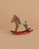 The Maileg Mouse Rocking Horse, featuring a brown body, green handle, and red rockers, rests on a beige surface—ideal for tiny mice or magnetic-handed figures to play and enjoy.