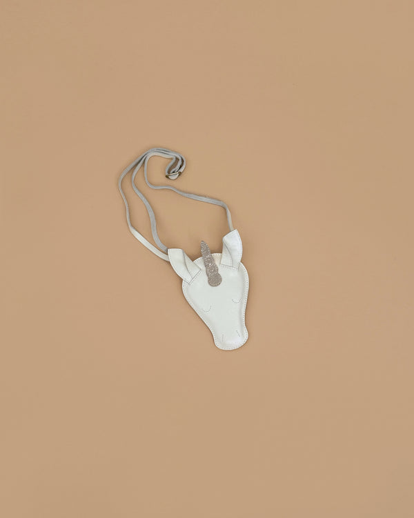 The Donsje Leather Blinc Purse - Unicorn is a small white purse with a silver glittery horn and adjustable leather strap, shown on a plain light brown background.