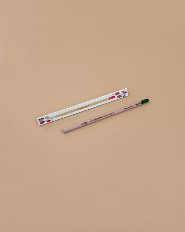 A Strawberry Plantable Pencil – Eco-Friendly with a green tip rests beside a white box adorned with red strawberries and leaves, both crafted from sustainable materials, set on a light brown background.