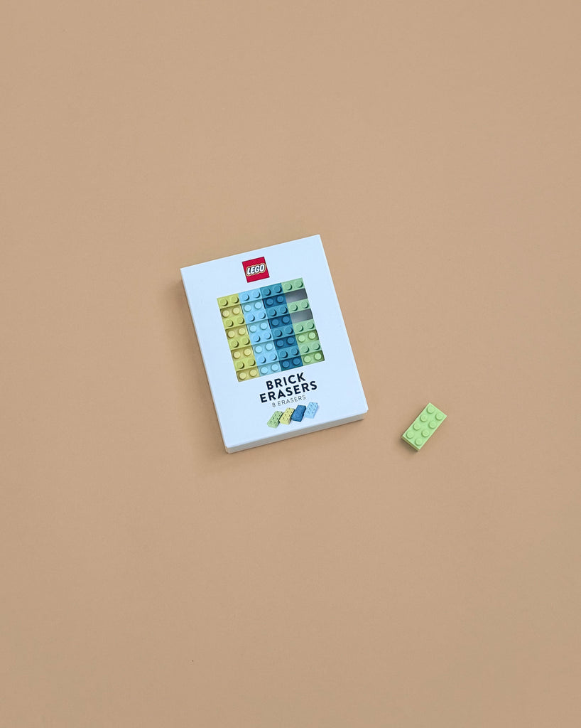 A white box labeled "Lego Brick Erasers" sits on a beige surface, featuring colorful erasers shaped like LEGO bricks. A green brick-shaped eraser lies beside the box, making this set a fun party favor for LEGO fans.