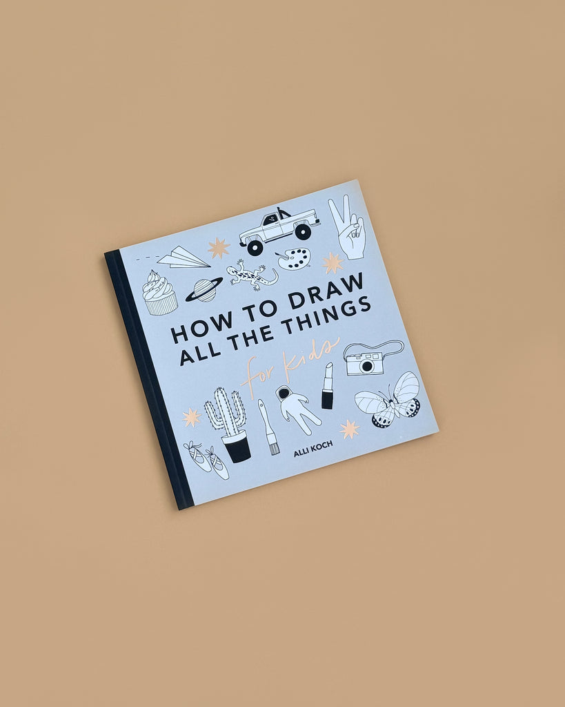 The book "All the Things: How To Draw Books For Kids" rests on a light brown surface, its cover featuring simple black-and-white line drawings—ideal for easy drawing lessons with items like a truck, pizza, camera, and plants.