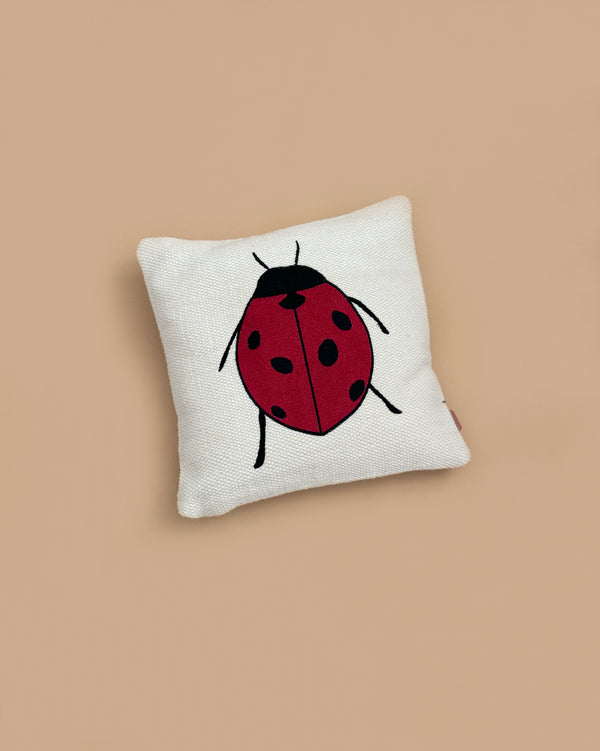 The Ferm Living Forest Embroidered Cushion - Ladybird is a white organic cotton cushion with a simple red ladybug featuring black spots and legs, set on a beige background.