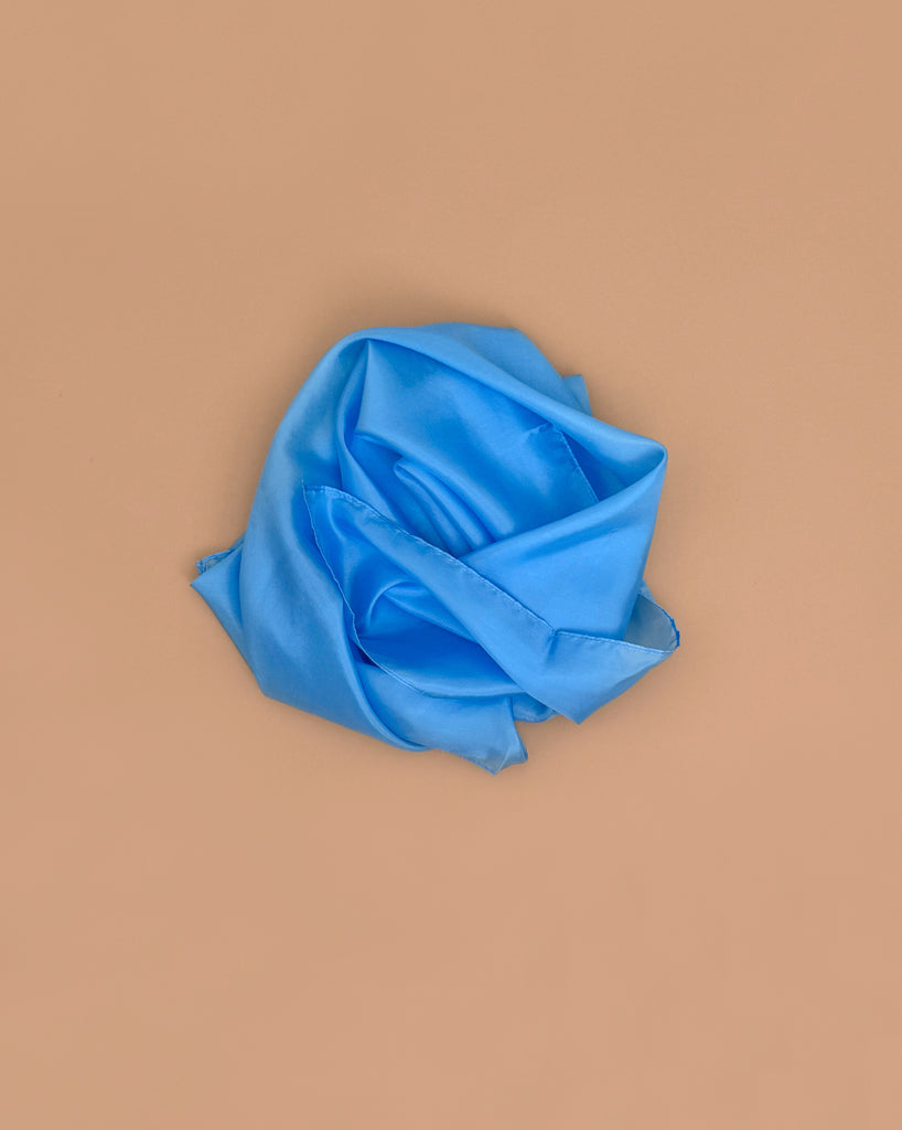 Sarah's Silks Solid Color Playsilk in Sky Blue is carefully folded to look like a rose and displayed on a plain beige background, highlighting its suitability for sensory play.