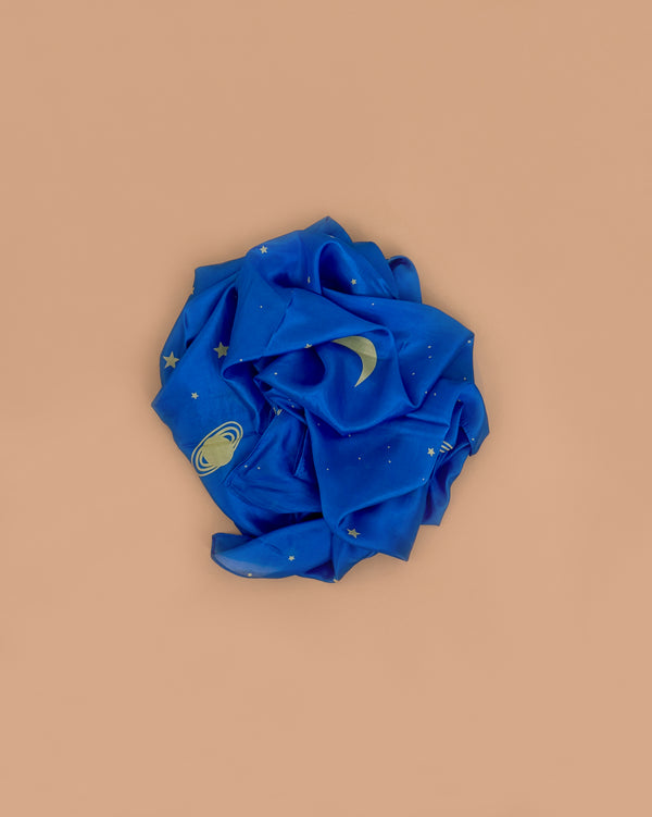 Sarah's Silks Giant Playsilk - Starry Night, a crumpled blue silk with gold stars, crescent moon, and ringed planet designs, rests on a plain beige background—ideal for imaginative play and open-ended fun.