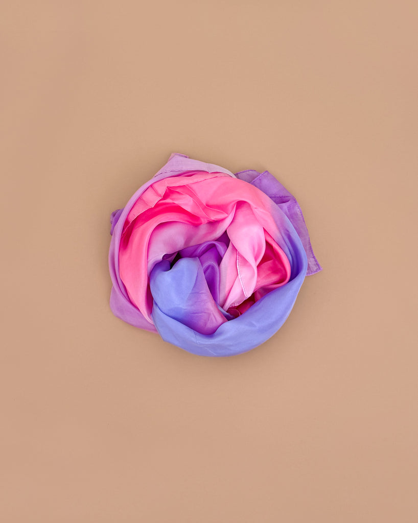 Sarah's Silks Giant Playsilk - Blossom, featuring pink, purple, and blue hues, is rolled into a circle on a beige background—ideal for open-ended play or enhancing your Giant Playsilks sensory play collection.
