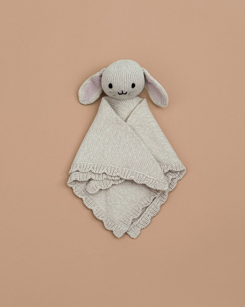 The Cuddle + Kind Organic Cotton Baby Bunny Lovey (Oatmeal), featuring floppy ears and embroidered facial details, is hand-knit and lies on a plain beige background.