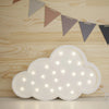 The Handmade Wooden Lamp - White Cloud, featuring glowing bulbs, rests on a pinewood-patterned surface against a light wood wall adorned with pastel and dark triangular pennants.