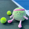 The Mon Ami - Tina Tennis Ball Plush Toy, shaped like a smiling tennis ball with a pink headband, striped arms and legs, and pink shoes, sits on a tennis court beside two real tennis balls.
