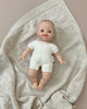 A Minikane Soft Body Doll (11") - Léo is lying on a textured beige blanket. The doll, perfect for imaginative playtime or as a bedtime companion, is wearing a simple white onesie and is positioned in the center of the knitted blanket, which has small holes in a repeating pattern.