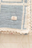 A close-up of the Organic Cotton Patchwork Baby Blanket - Spring Bloom, showing its blue and cream crochet trim with a scalloped edge and a "REVIVAL" fabric tag on the corner, folded on a light quilted surface.