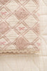 A close-up of the Organic Cotton Baby Patchwork Blanket - Pink Bows, featuring a diamond quilted pattern, pink and cream tones, embroidered bows, and scalloped edges, displayed on a matching textured surface. Ships in one week.