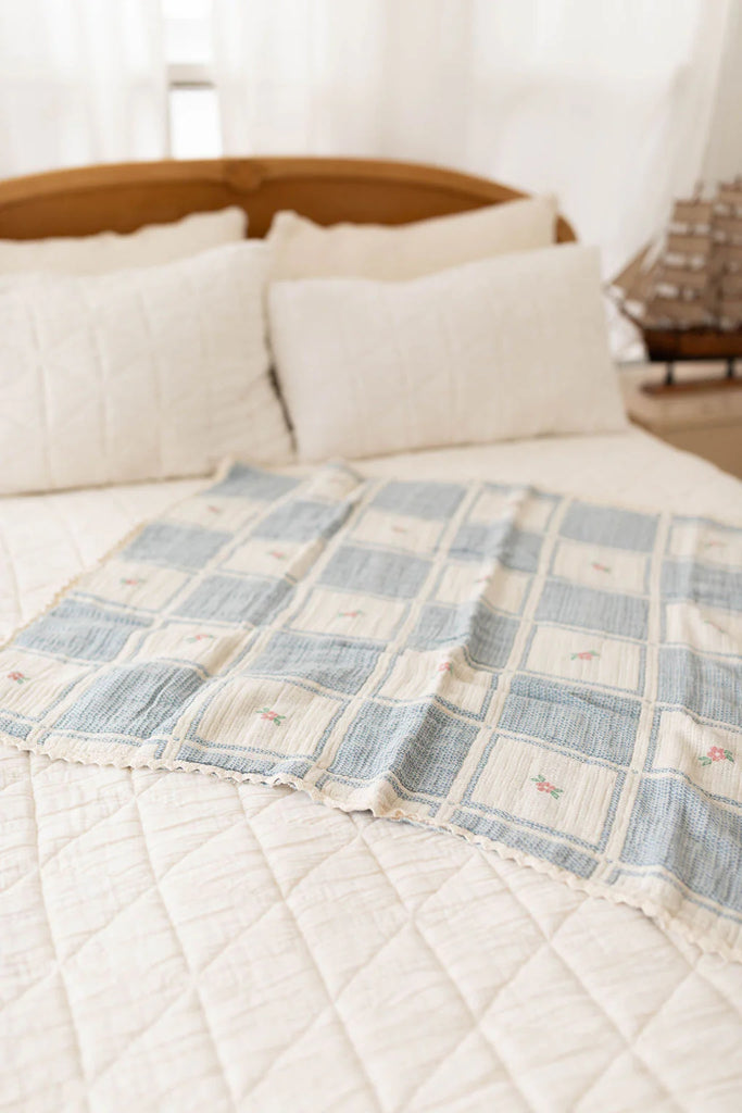 The Organic Cotton Patchwork Baby Blanket - Spring Bloom, featuring light blue, cream checks, and pink floral details, is displayed on a neatly made bed with white quilted bedding and large pillows; a model ship is in the background.