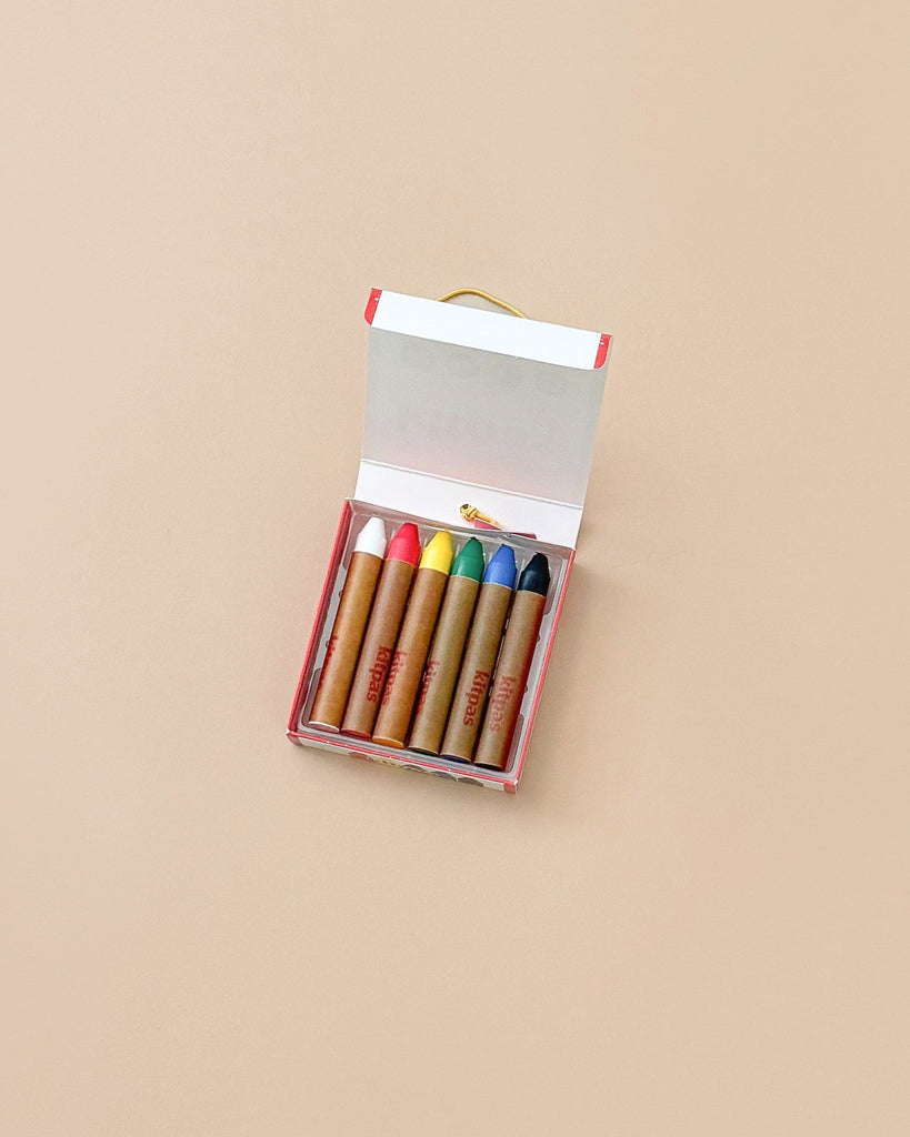 An open box of Kitpas Crayons - 6 Colors sits on a plain beige background. The six thick, non-toxic crayons have brown paper wrappers and are in a white box with red edges.