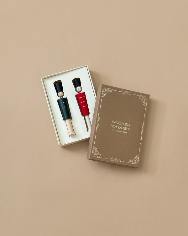 A box labeled "Wooden Soldiers," elegantly adorned with gold patterns, is open on a beige surface, revealing two hand-painted wooden soldier figurines—one dressed in a red uniform and the other in a black uniform.
